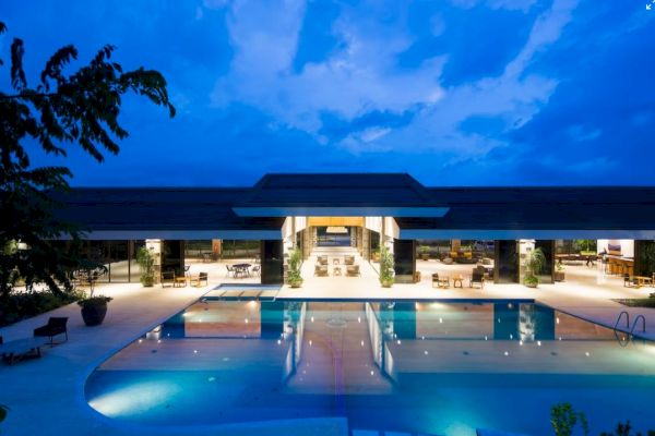 A luxurious modern building with a large pool in front, lit up in the evening under a blue sky.