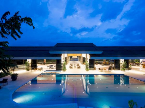 A luxurious modern building with a large pool in front, lit up in the evening under a blue sky.