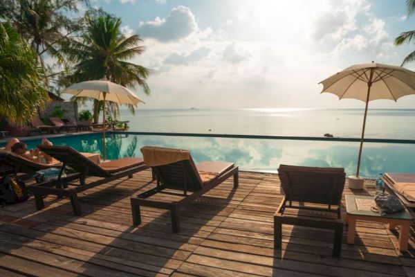 A serene pool setting with lounge chairs, umbrellas, palm trees, and a tranquil ocean view under a cloudy sky.