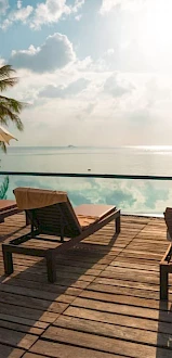 Poolside scene with lounge chairs, umbrellas, and a calm water view; palm trees and sunlit sky create a tranquil atmosphere.
