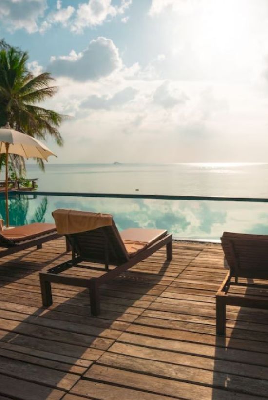 Poolside scene with loungers, umbrellas, and palm trees overlooking a serene ocean under a partly cloudy sky.