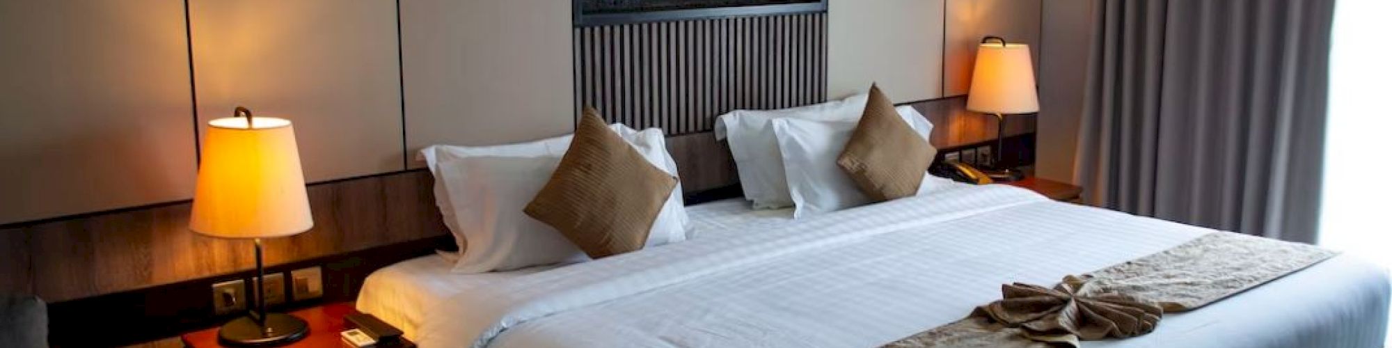 A modern hotel room with a neatly made bed, two bedside lamps, decorative pillows, and curtains, creating a cozy and inviting atmosphere.
