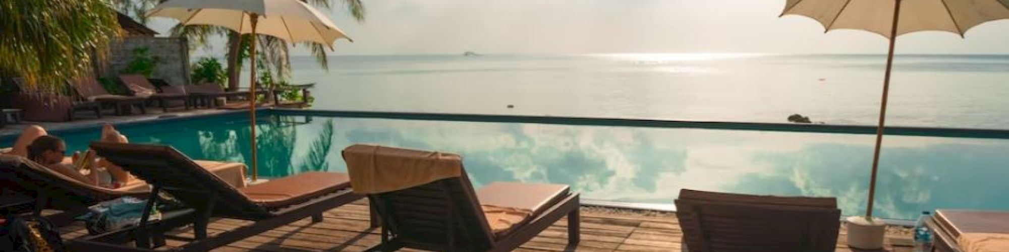 A serene poolside scene with lounge chairs, umbrellas, palm trees, and an ocean view under a partly cloudy sky.