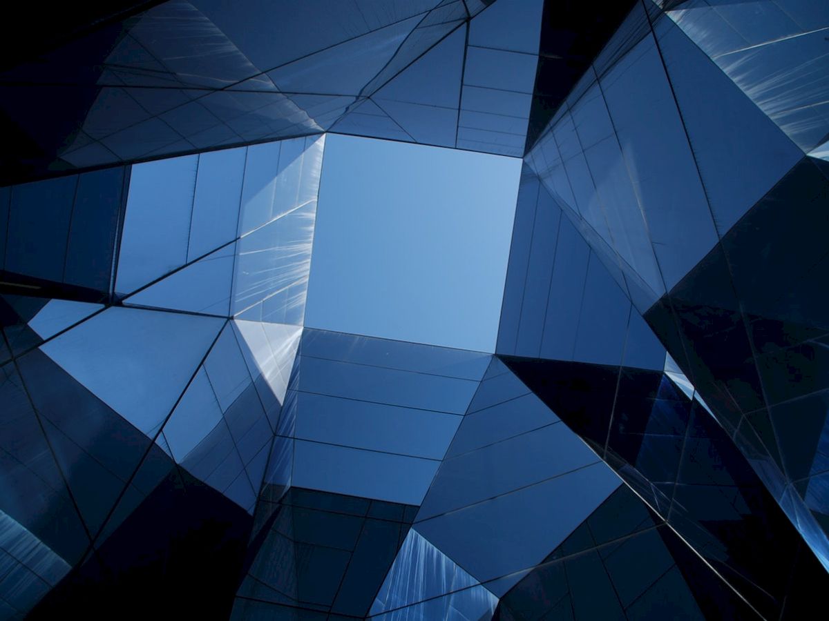 Looking up at a geometric structure of glass panels, reflecting the sky and surrounding environment, creating a kaleidoscope effect.