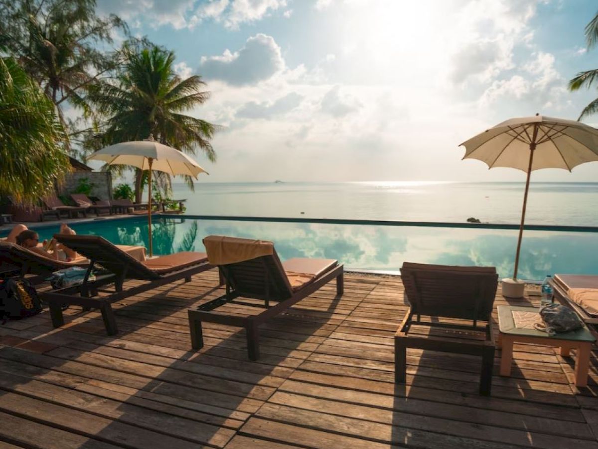 A serene poolside setting with lounge chairs, umbrellas, and palm trees overlooking a calm ocean under a partially cloudy sky.