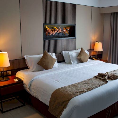 A well-lit hotel room features a large bed with pillows and throws, two lamps on side tables, and a decorative wall piece.