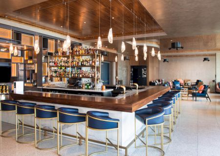 A modern bar with a long counter, blue chairs, hanging lights, and a well-stocked back bar. The setting appears stylish and inviting.