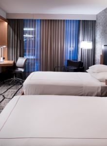 The image shows a modern hotel room with two beds, a desk, a chair, wall-mounted TV, and a window with curtains, creating a cozy atmosphere.
