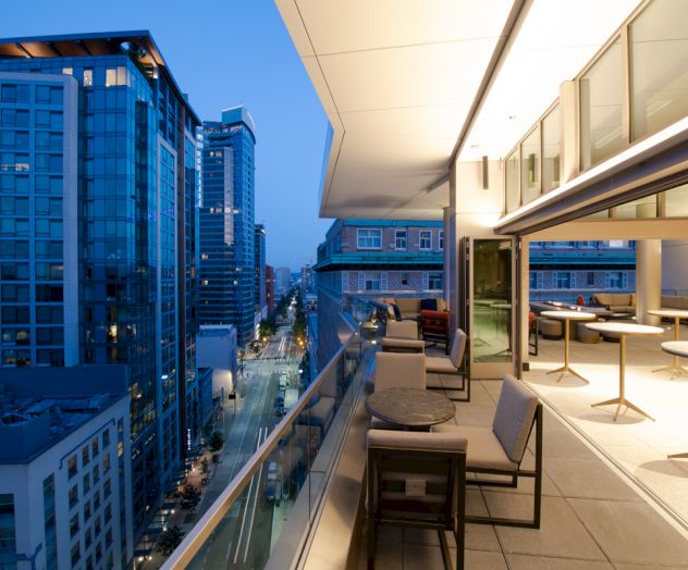 A modern balcony overlooks a city street, with tall glass buildings and urban lights visible during dusk.