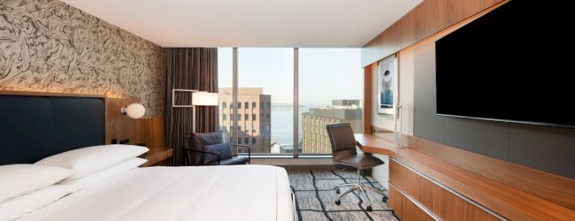 A modern hotel room with a large bed, desk, chair, and a flat-screen TV. The room has a view of buildings through a wide window.