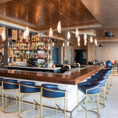 A modern bar with blue stools, pendant lights, and a well-stocked liquor shelf. The seating area features a stylish design.