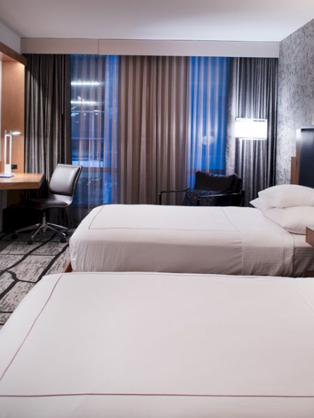 The image shows a modern hotel room with two beds, a TV, a desk, a chair, and stylish lighting.