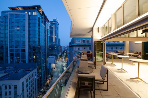 A modern cityscape with tall buildings is seen from a balcony at dusk. The balcony has seating and tables, offering an inviting atmosphere.