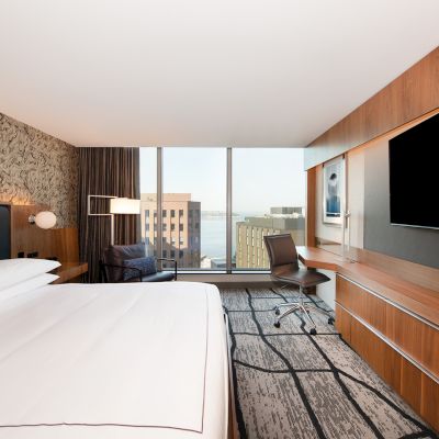 A modern hotel room with a large bed, desk, chair, and flat-screen TV. It has a city view through a large window.