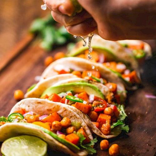 Tacos filled with chickpeas, avocado, and vegetables on a wooden board, with lime being squeezed over them.
