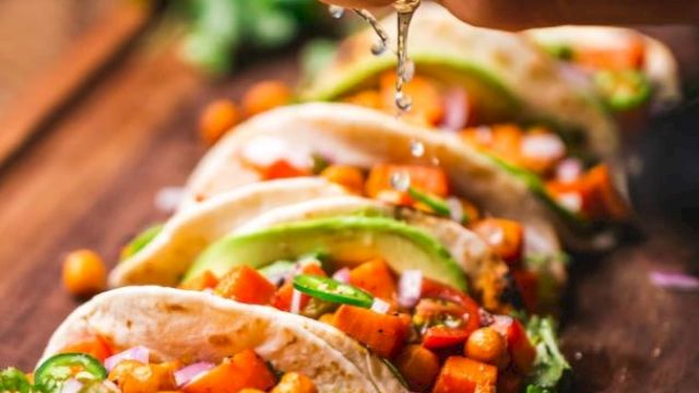 Tacos filled with chickpeas, avocado, and vegetables on a wooden board, with lime being squeezed over them.