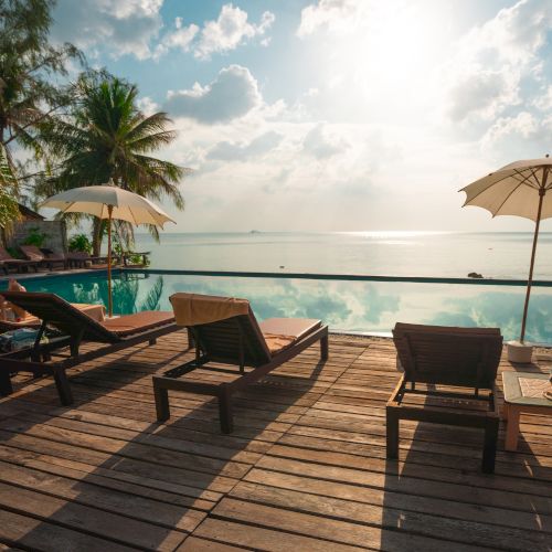 A serene wooden deck with lounge chairs, umbrellas, overlooking an infinity pool and ocean under a bright sky and palm trees.