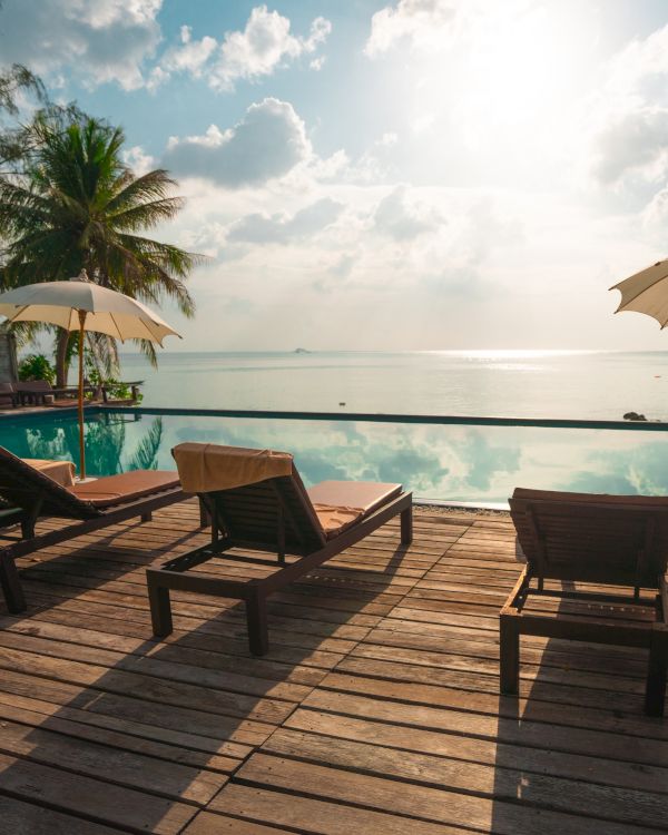 A serene wooden deck with lounge chairs, umbrellas, overlooking an infinity pool and ocean under a bright sky and palm trees.