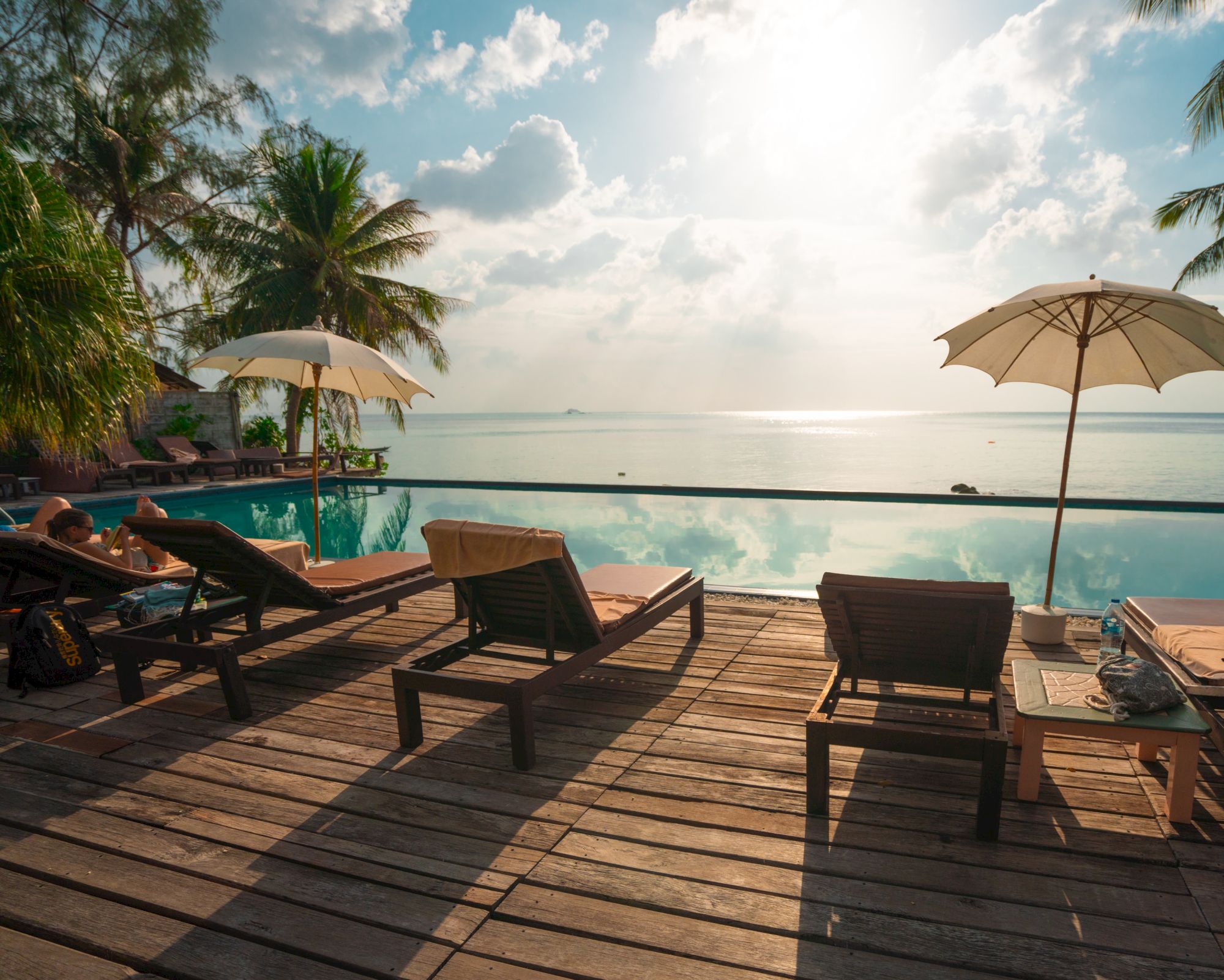Lounge chairs on a wooden deck overlook a calm infinity pool, with palm trees and umbrellas under a sunny, partly cloudy sky by the sea.