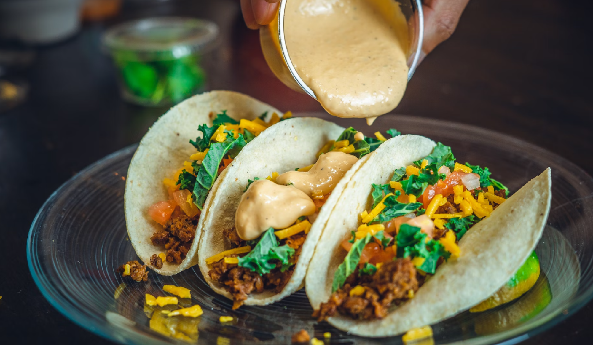 Three tacos on a plate filled with ground meat, cheese, kale, and tomatoes, being topped with creamy sauce.