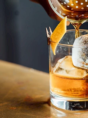 A cocktail is being poured into a glass with ice and a lemon twist garnish on a wooden surface.