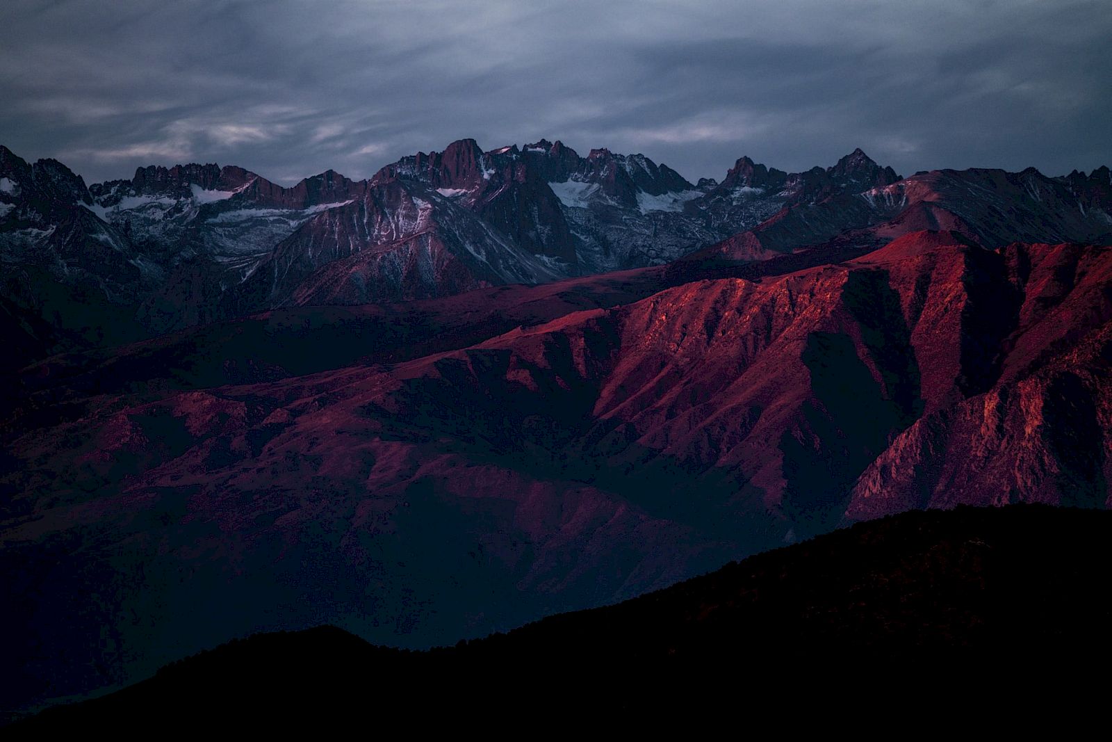 A mountain range with snow-capped peaks, bathed in a red-hued light under a cloudy sky.