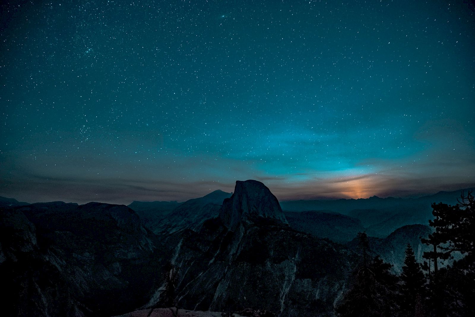 A night sky filled with stars over a silhouette of mountains and trees, with a glow from the horizon suggesting a rising or setting sun.