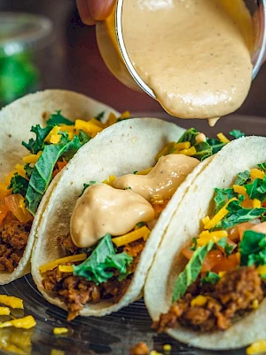 Three tacos filled with ground meat, lettuce, tomatoes, and cheese, topped with sauce being poured over them.