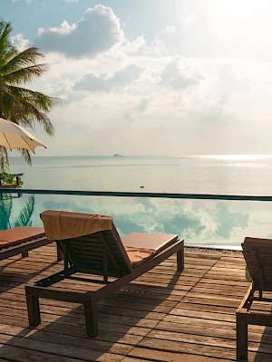 A serene poolside scene with lounge chairs, parasols, and palm trees overlooking a calm sea under a partly cloudy sky.
