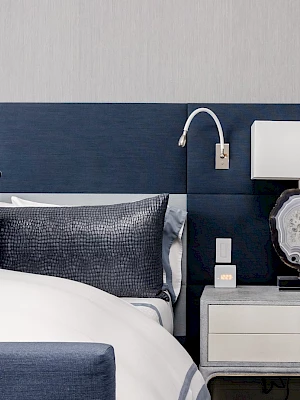 A modern bedroom with a blue headboard, grey pillows, a white lamp on a nightstand, and a textured wall backdrop.