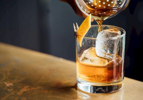 A cocktail is being poured into a glass filled with ice and garnished with a twist of citrus peel pinned to the rim.