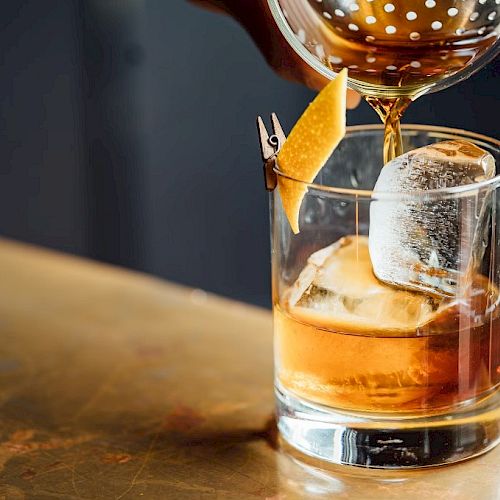 A cocktail is being poured into a glass filled with ice and garnished with a twist of citrus peel pinned to the rim.