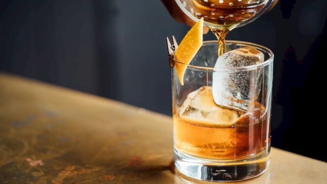 A cocktail is being poured into a glass filled with ice and garnished with a twist of citrus peel pinned to the rim.