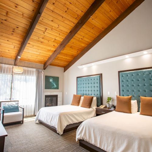 The image shows a cozy bedroom with two beds, wood-beamed ceiling, textured blue headboards, and natural light from a large window.