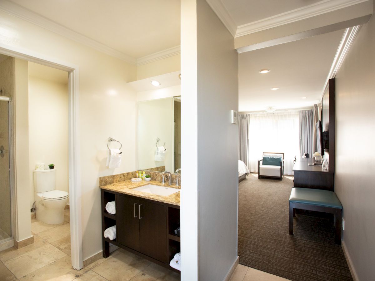 The image shows a hotel room with a bathroom on the left and a bedroom area on the right, featuring a bed and seating.