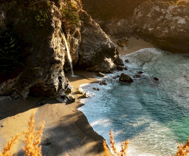 A scenic coastal view with a waterfall cascading onto a secluded beach, surrounded by rocky cliffs and lush greenery.