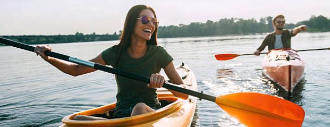 Two people are kayaking on a calm lake, smiling at each other under the sun, enjoying the outdoor activity and nature.