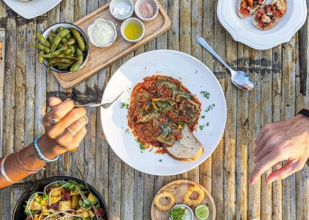 People's hands reaching for dishes on a bamboo table, featuring pasta, salad, appetizers, drinks, and condiments.