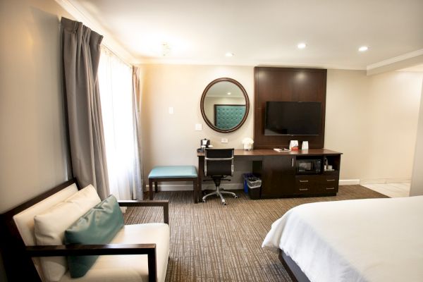 A modern hotel room with a bed, armchair, desk, TV, mirror, and minimal decor.