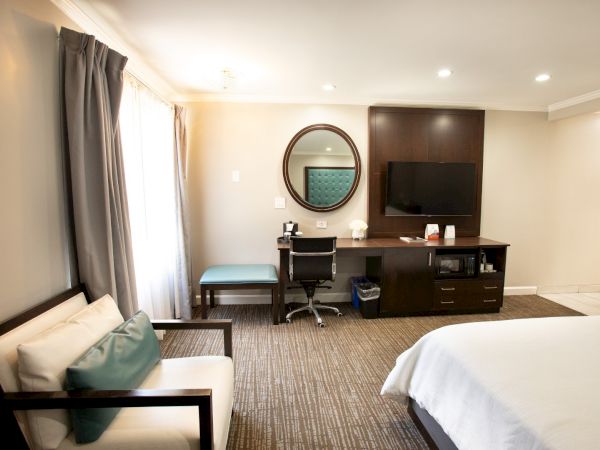 A modern hotel room with a bed, armchair, desk, TV, mirror, and minimal decor.