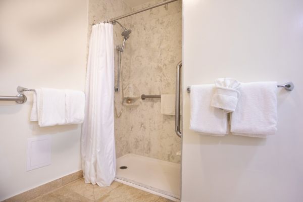 The image shows a bathroom with an accessible walk-in shower, grab bars, a white curtain, and two folded towels on racks.