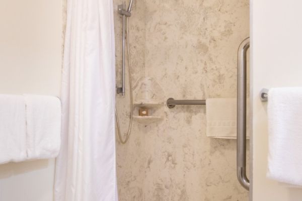 The image shows a bathroom with a walk-in shower, a white curtain, and a grab bar. There are towels hanging on the left wall.