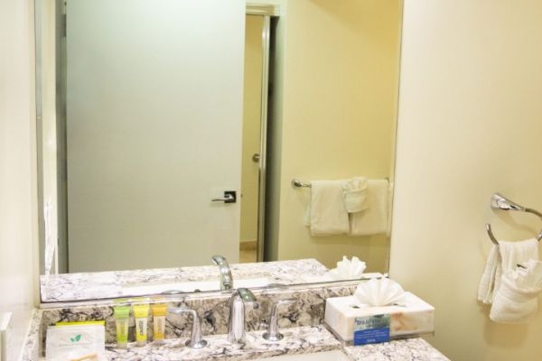 A bathroom with a marble countertop, sink, toiletries, a wall-mounted hairdryer, large mirror, and towel hanging on a rack.