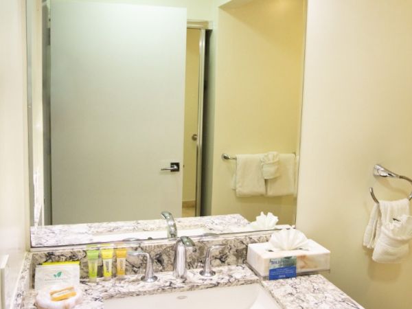 A bathroom with a marble countertop, sink, toiletries, a wall-mounted hairdryer, large mirror, and towel hanging on a rack.