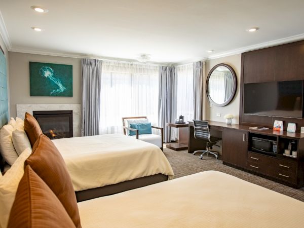 A hotel room with two beds, a TV, a desk, a mirror, a fireplace, and a seating area by the window.