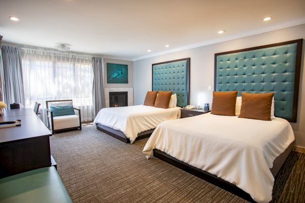 This image shows a hotel room with two beds, teal headboards, desk, chairs, and a window with curtains, creating a cozy atmosphere.