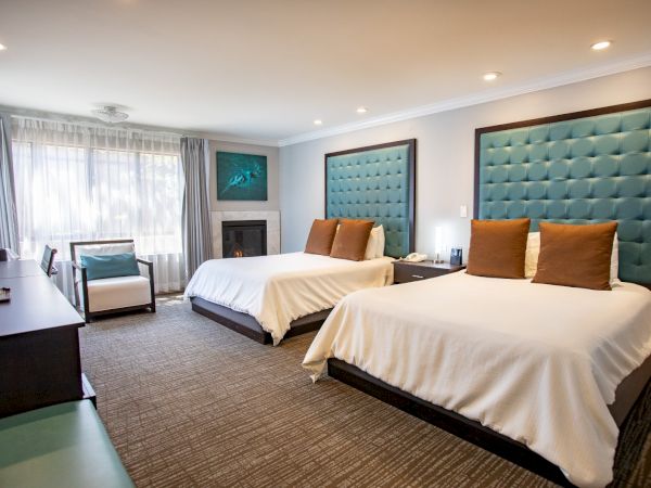 This image shows a hotel room with two beds, teal headboards, desk, chairs, and a window with curtains, creating a cozy atmosphere.