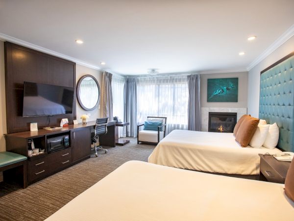 The image shows a hotel room with two beds, a TV, a fireplace, seating, a desk, and window curtains, in a softly lit modern setting.