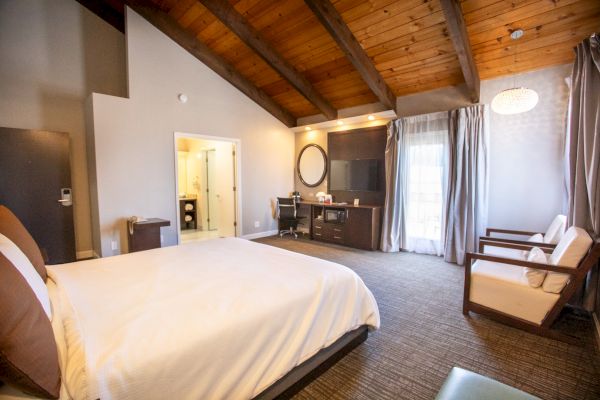 The image shows a spacious hotel room with a large bed, wooden ceiling, chairs, a desk, and an en-suite bathroom.