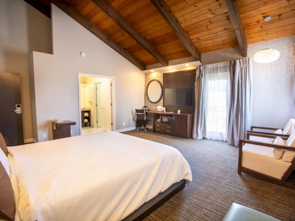 The image shows a spacious hotel room with a large bed, wooden ceiling, chairs, a desk, and an en-suite bathroom.